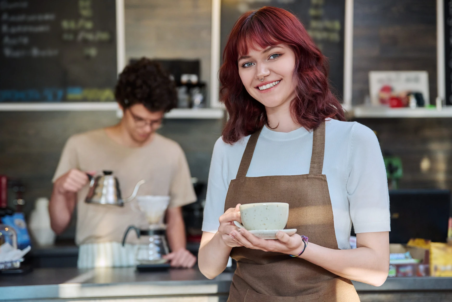 Portrait of young smiling female waitress in an apron holding cup of coffee, standing in front of counter in coffee shop. Food service occupation, part time job for students, staff, work concept