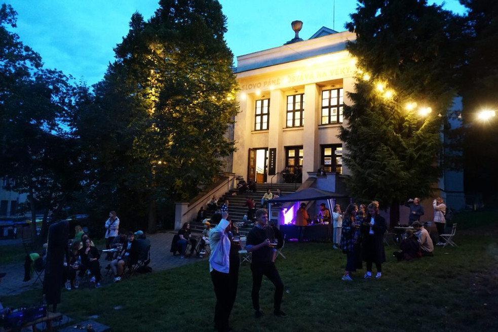 Pohled na budovu Komunitního prostoru Smíchov. Atmosféra letního večera. Lidé sedí na zahrádce venku před budovou. Nad lidmi svítí světýlka.